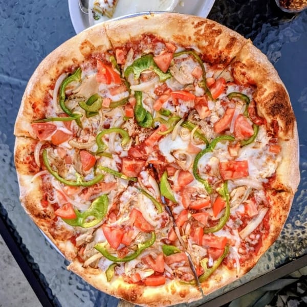 A whole Italian pizza with green peppers, onions, tomatoes, and chicken on a glass table.