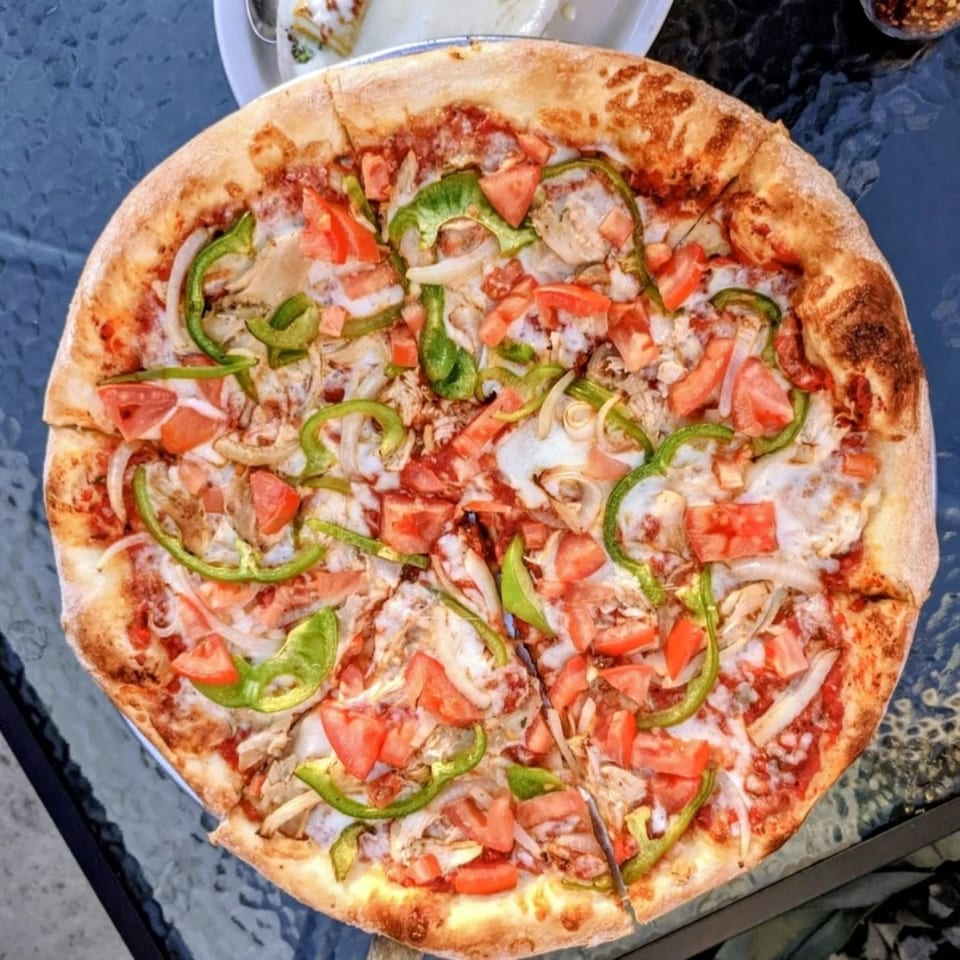 A whole Italian pizza with green peppers, onions, tomatoes, and chicken on a glass table.