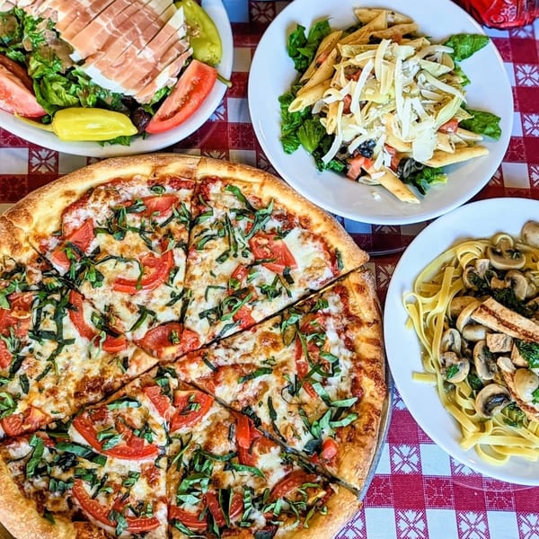 A full Italian meal with a large pizza, two pasta dishes, and a salad on a red and white checkered tablecloth.