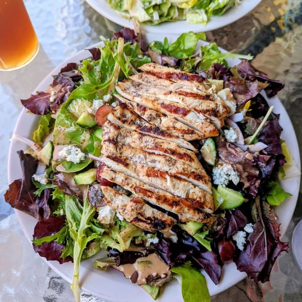 Grilled chicken salad with mixed greens, avocado, red onion, and feta cheese on a white plate.