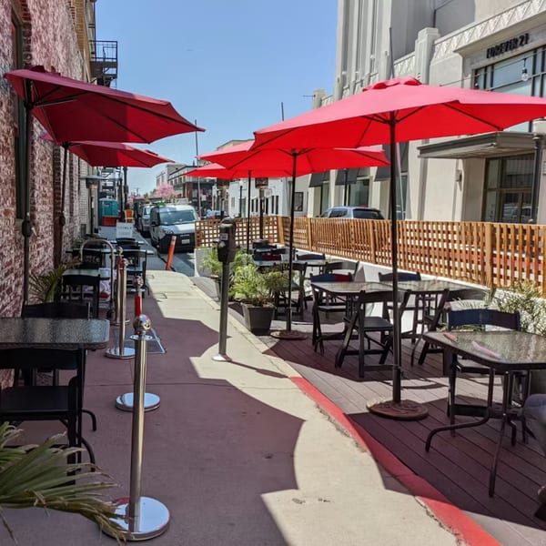 Outdoor patio dining area with red umbrellas, tables, and chairs along a brick building on a sunny day.
